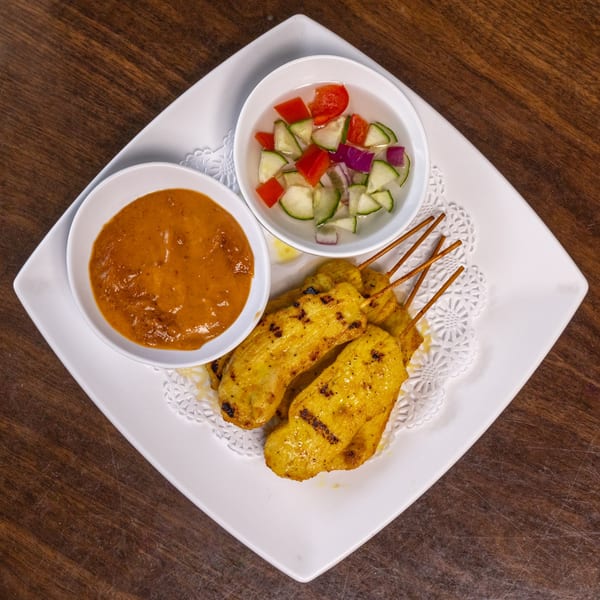 Thai chicken satay skewers with peanut sauce and cucumber salad on a white plate.